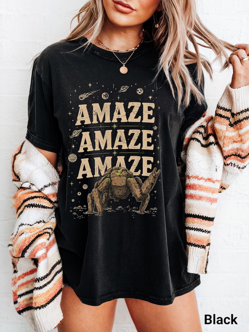 May include: Black t-shirt with the word "AMAZE" repeated three times in a tan font. A brown creature graphic is below the text, with planets and stars in the background. The word "Black" is at the bottom right.