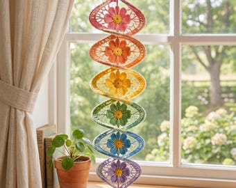Può includere: Una decorazione a spirale sospesa con sei motivi floreali all'uncinetto in un gradiente verticale arcobaleno. Ogni sezione contiene un fiore con un centro giallo e petali nei colori rosa, arancione, giallo, verde, blu e viola, uniti da filato.