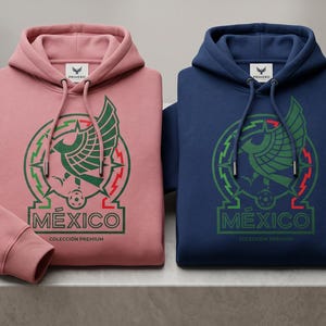 Pu&ograve; includere: Due felpe con cappuccio, una rosa polvere e una blu navy, ciascuna con la grafica di un'aquila stilizzata, la parola MEXICO e il testo COLECCI&Oacute;N PREMIUM. Entrambi i capi presentano cordini abbinati e un'etichetta del marchio sul colletto.