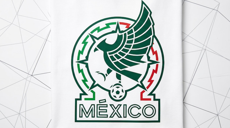 Pu&ograve; includere: Una t-shirt bianca con un logo grafico raffigurante un'aquila verde stilizzata con le ali spiegate, posizionata sopra un pallone da calcio. Il design &egrave; racchiuso in una cornice circolare con accenti rossi e verdi e la parola M&Eacute;XICO in grassetto.