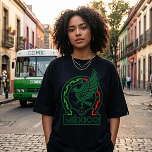 Pu&ograve; includere: Una t-shirt nera a maniche corte con una stampa grafica di un emblema d'aquila verde con accenti rossi e bianchi e il testo MEXICO centrato sul petto. Il design include un bordo circolare e un'icona di un pallone da calcio.