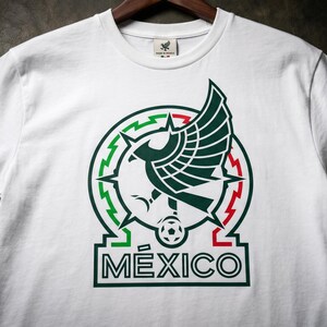 Pu&ograve; includere: Una t-shirt bianca a maniche corte con una grafica centrale raffigurante un'aquila stilizzata in verde scuro, posizionata sopra un pallone da calcio. Il design &egrave; incorniciato da un bordo circolare verde, bianco e rosso, con la scritta M&Eacute;XICO in verde scuro.