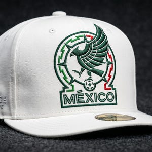 Pu&ograve; includere: Un cappellino da baseball bianco con un emblema a forma di aquila verde ricamato sopra la scritta M&Eacute;XICO. Il design include un pallone da calcio all'interno di un cerchio. Il lato del cappellino mostra la scritta ricamata HERITAGE COLLECTIVE.