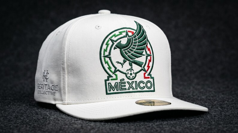 Pu&ograve; includere: Un cappellino da baseball bianco con un emblema a forma di aquila verde ricamato sopra la scritta M&Eacute;XICO. Il design include un pallone da calcio all'interno di un cerchio. Il lato del cappellino mostra la scritta ricamata HERITAGE COLLECTIVE.