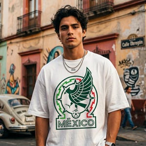 Pu&ograve; includere: Una t-shirt bianca a maniche corte con una grande stampa grafica sul petto. Il design mostra un emblema stilizzato di un'aquila verde con accenti rossi e la parola M&Eacute;XICO stampata in grassetto con lettere maiuscole verdi sotto la grafica.