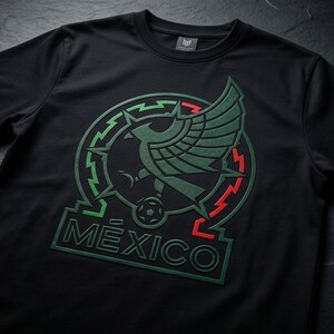 Pu&ograve; includere: Una t-shirt nera a maniche corte con una grande grafica sul petto. Il design include un'aquila stilizzata in verde scuro, un pallone da calcio e la parola M&Eacute;XICO in lettere in grassetto, incorniciata da un bordo circolare con accenti verdi e rossi.