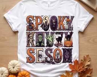 Può includere: Una t-shirt bianca a maniche corte con una grande stampa grafica con il testo SPOOKY HOUTY SESON. Il design include illustrazioni di una strega, un lupo mannaro, un vampiro, uno scheletro, una mummia, Frankenstein, un fantasma, un gatto nero, una zucca e un ragno.