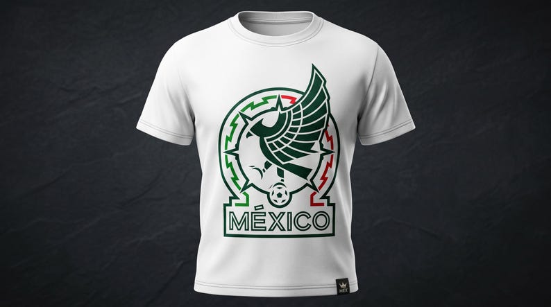 Pu&ograve; includere: Una t-shirt bianca a maniche corte con una grafica centrale raffigurante un'aquila stilizzata con le ali spiegate, un pallone da calcio e la scritta M&Eacute;XICO in verde scuro e rosso. Il design &egrave; circolare con accenti geometrici.
