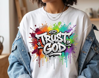 Può includere: Una t-shirt bianca a girocollo con un vivace design a schizzi di vernice multicolore come sfondo per il testo "TRUST GOD" in stile graffiti, con lettere bianche in grassetto, contorni neri e una piccola aureola sopra la parola trust.
