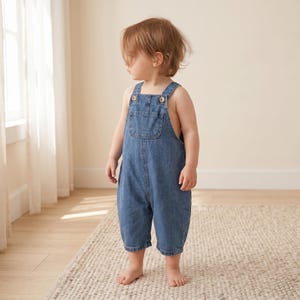 May include: A toddler in a blue denim dungaree jumpsuit with wooden button fastenings. The sleeveless jumpsuit features a front pocket and is worn barefoot. The child is standing on a light-coloured rug.