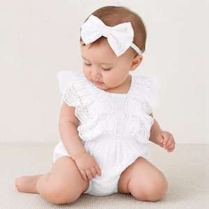 May include: A white sleeveless romper featuring a textured fabric and large lace ruffles along the bodice. The garment is paired with a matching white lace bow headband. The design includes a gathered waistline and a simple, clean aesthetic.