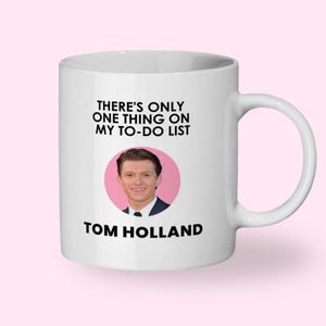 May include: White ceramic mug with a handle featuring the text "THERE'S ONLY ONE THING ON MY TO-DO LIST" above a photo of Tom Holland in a pink circle, with "TOM HOLLAND" printed below.