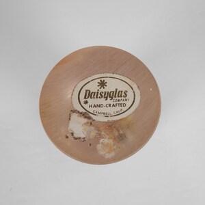 May include: A close-up of a round, wooden bottle stopper with a gold label that reads "Daisyglas Company * Hand-Crafted Campbell Calif."