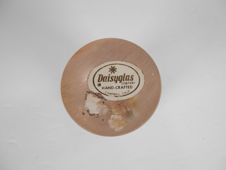May include: A close-up of a round, wooden bottle stopper with a gold label that reads "Daisyglas Company * Hand-Crafted Campbell Calif."
