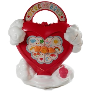 May include: A red heart-shaped plastic toy case with a white cloud base and side accents. The handle features Care Bears characters. The front displays a circular spinner with icons including a rainbow, sun, and cupcake, plus a small pink cupcake figure.