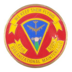 May include: A round, gold-colored pin with a red border and the text "WE KEEP THEM FLYING" in yellow. The center features a blue triangle with a gold airplane and a hand holding tools. The text "42nd ORGANIZATIONAL MAINTENANCE SOON" is also present.