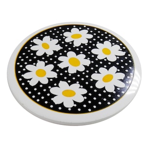 Puede incluir: Un posavasos redondo con borde blanco. El centro presenta un fondo negro con lunares blancos y seis margaritas blancas con centros amarillos. El posavasos probablemente está hecho de cerámica o un material similar.