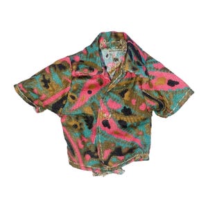 May include: A short-sleeved button-down shirt with a colorful pattern. The shirt features a design with pink, teal, brown, and black shapes. The collar and button details are visible. The shirt is likely made of a lightweight fabric.