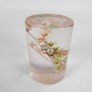 May include: A clear glass cylinder filled with a bouquet of dried flowers. The flowers are white, green, and brown. The cylinder is sitting on a white surface.