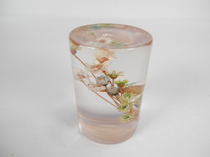 May include: A clear glass cylinder filled with a bouquet of dried flowers. The flowers are white, green, and brown. The cylinder is sitting on a white surface.