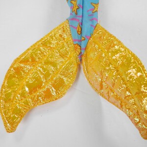 May include: A pair of yellow mermaid tail fins with a shimmery finish. The fins are made of fabric and have a textured surface.