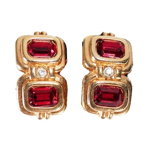 May include: A pair of gold-toned clip-on earrings. Each earring features two rectangular, deep red gemstones and a small, round, clear gemstone. The earrings have a geometric design with a vintage aesthetic.