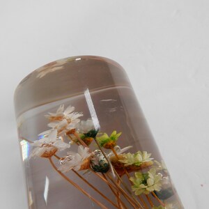 May include: A clear glass cylinder filled with dried white and green flowers. The flowers are arranged in a bouquet and are suspended in the clear liquid.