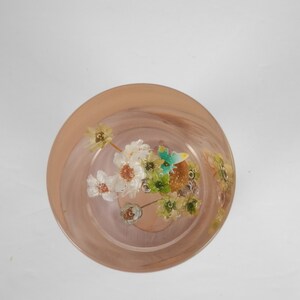 May include: A clear glass bowl with a pink rim filled with dried flowers and leaves. The flowers are white and yellow, and the leaves are green.