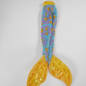 May include: A blue and yellow mermaid tail with a pink and yellow seashell pattern. The tail is made of fabric and has a shiny gold finish.