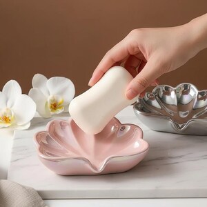 May include: A pink, shell-shaped soap dish with a pearlescent finish. A white bar of soap is being placed in the dish. A second silver-colored shell-shaped dish is visible. White orchids are in the background. The dish is on a white marble surface.