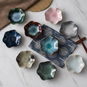 May include: A collection of nine small, ceramic, flower-shaped dishes in various colors, including blue, green, pink, and gray. Some have a gold rim. The dishes are arranged on a marble surface with a blue patterned cloth and chopsticks.