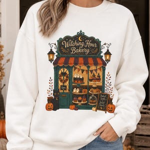 May include: A white crewneck sweatshirt featuring a graphic illustration of a bakery storefront labeled "Witching Hour Bakery." The design includes a black cat, pumpkins, hanging lanterns, and a display window filled with cakes and pastries.