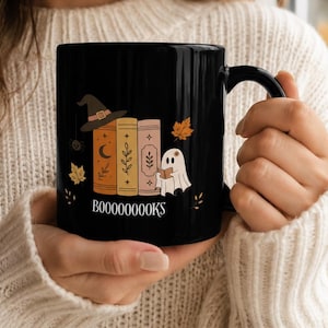 Op de afbeelding: Een zwarte keramische mok met een afbeelding van drie gestapelde boeken, een heksenhoed en een klein spookje dat een boek leest. Het ontwerp bevat herfstbladeren en de tekst BOOOOOOOOKS in het wit onderaan de illustratie.