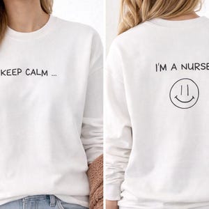 以下が含まれることがあります： 前面に「KEEP CALM ...」、背面にスマイリーフェイス付きの「I'M A NURSE」と書かれた白いスウェットシャツ。テキストとスマイリーは黒色です。