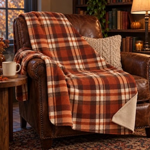 May include: A soft throw blanket featuring a plaid pattern in shades of burnt orange, deep brown, and cream. The blanket has a folded corner revealing a solid, light beige fleece-like underside, draped over a brown leather armchair.