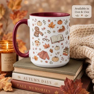 May include: A white ceramic mug with a deep red handle and interior, featuring a repeating pattern of autumn-themed illustrations including pumpkins, a plaid scarf, an open book, a cardigan, and a slice of pie. Text on the mug reads Available in 11oz & 15oz.