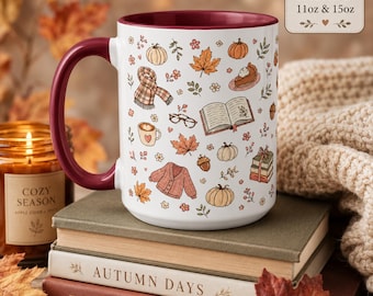 May include: A white ceramic mug with a deep red handle and interior, featuring a repeating pattern of autumn-themed illustrations including pumpkins, a plaid scarf, an open book, a cardigan, and a slice of pie. Text on the mug reads Available in 11oz & 15oz.
