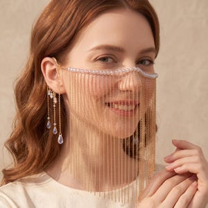 May include: A gold-toned chain face mask featuring a horizontal strand of small white pearls across the bridge of the nose. Numerous delicate gold chains hang vertically from the pearl strand, and additional chains with pearl and teardrop accents.