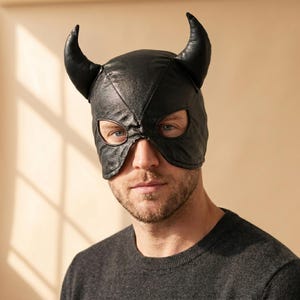 May include: A black leather mask featuring two prominent, curved horns extending from the top. The mask covers the upper portion of the face, including the forehead and temples, with two distinct eye openings and a pointed center seam design.
