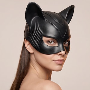 May include: Black leather cat mask with pointed ears and eye cutouts. The mask has a sleek, sculpted design with textured details. The mask covers the eyes and nose, and is worn on the face.