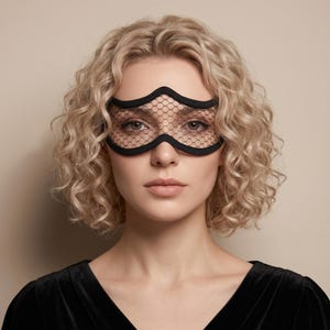 May include: A black eye mask with a decorative wavy top edge and a mesh design over the eyes. The mask is worn by a person with curly blonde hair and a black top.