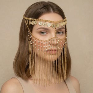May include: A gold-coloured headpiece with a face veil. The headpiece has a chain that goes around the head, decorated with small circular pendants. The face veil is made of chains and dangling crystals, creating an elegant and decorative appearance.
