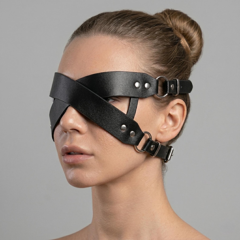 May include: Black leather blindfold with crisscross design. The blindfold features metal hardware, including rivets and adjustable buckles. The straps wrap around the head and ears. The product is designed for sensory play.