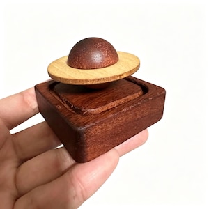 Pode incluir: Um brinquedo de quebra-cabeça de madeira com uma base quadrada castanho-escura com um centro rebaixado. Uma esfera castanho-escura menor está posicionada no meio, rodeada por um anel de madeira claro e plano colocado horizontalmente.