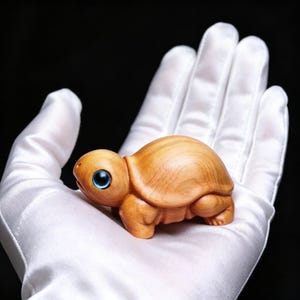 May include: A small, wooden turtle figurine with a light brown finish. The turtle has detailed carving and bright blue eyes. The figurine is held in a white gloved hand against a black background. The turtle is a decorative item.
