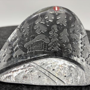 Può includere: Una scultura in vetro trasparente, triangolare, con dettagli incisi di una scena invernale. Il design include una baita, alberi e una strada. La scultura è contrassegnata "Made in Finland".