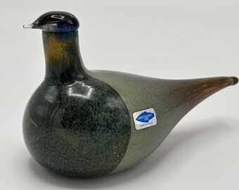 May include: A glass bird figurine featuring a dark blue head, a mottled dark green chest, and a light gray body that transitions into a brown tail. A blue and white sticker on the side reads Noutajarvi 1793 Made in Finland.