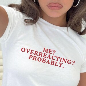 Può includere: T-shirt bianco con la scritta rossa "ME? OVERREACTING? PROBABLY." La maglietta è a girocollo e sembra essere realizzata in un materiale morbido. Il testo è centrato sul davanti della maglietta.