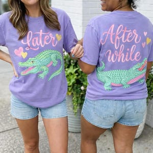May include: Two lavender t-shirts with a cartoon alligator design. One shirt says "later" on the front and "After While" on the back. The alligator is green with pink accents. The shirts are paired with denim shorts.