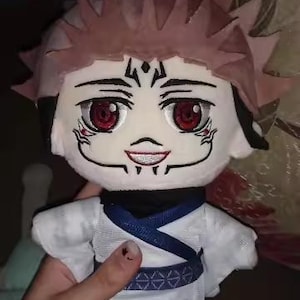 May include: A plush doll featuring a character with spiky pink hair, red eyes, and facial markings. The doll wears a white outfit with a blue sash and black shoes. The doll is held by a hand.